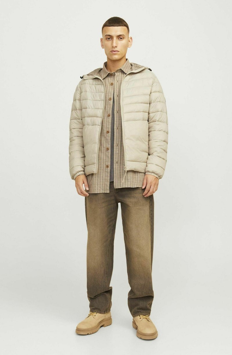 Doudoune beige avec capuche, dotée de coutures horizontales et d'une fermeture éclair à l'avant. Portée par-dessus une chemise à carreaux et un pantalon marron.