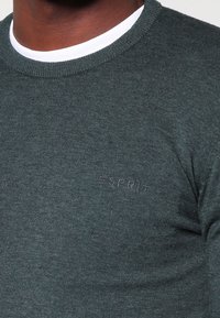 Czarnoszary sweter dzianinowy z subtelnym logo "ESPRIT" na klatce piersiowej, noszony na białej koszulce z okrągłym dekoltem.