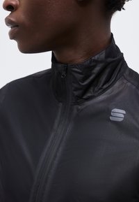 Közeli kép egy személyről, aki fekete, cipzáros, magas nyakú dzsekit visel, melynek mellkasán fényvisszaverő logó látható.