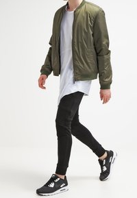 Olivově zelená bomber bunda se zipem vpředu, nošená přes dlouhou bílou košili. Černé skinny kalhoty a černé sportovní boty dokončují vzhled.