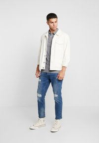 Lett jakke over en grå polo, kombinert med slitte blå jeans og hvite høyhælte joggesko. Enkelt design, avslappet passform.