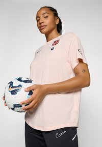 Roza sportska majica s detaljima emblema i logotipa, uparena s tamnim hlačama. Model drži šareni loptičasti nogomet, prikazujući aktivnu odjeću.