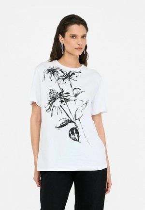 Weißes Baumwoll-T-Shirt mit einem schwarzen Blumenmuster. Kurze Ärmel und runder Halsausschnitt. Kombiniert mit schwarzen Hosen.