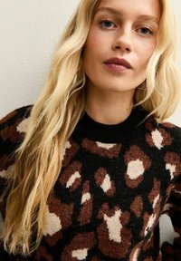 Blonde vrouw met lang golvend haar, gekleed in een zwart, bruin en beige trui met dierenprint, staande tegen een effen lichte muur.