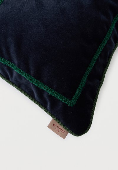 Coussin en velours bleu foncé avec une bordure brodée verte et une étiquette beige portant la mention "ETRO HOME" sur une surface blanche.