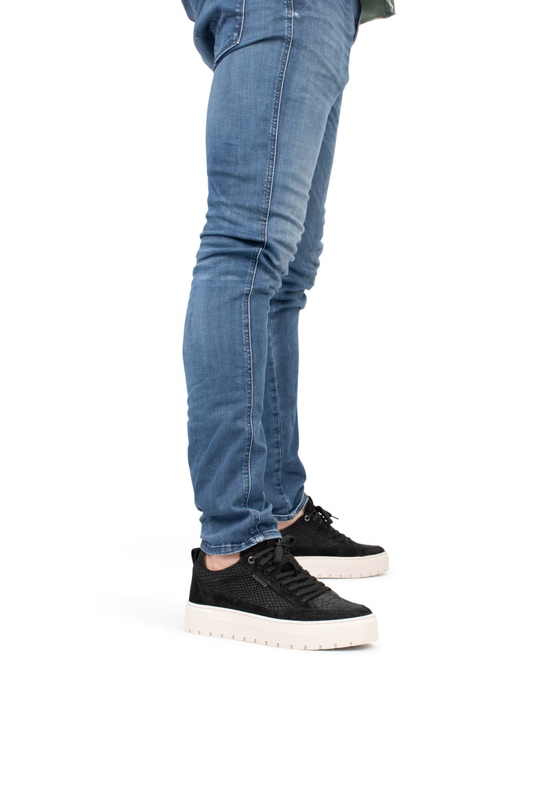 Zapatillas negras con parte superior texturizada, con una suela blanca suave. Combinadas con unos jeans ajustados de denim azul.