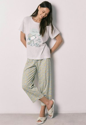 Vrouw glimlacht, draagt een lichtgrijs Snoopy T-shirt met opdruk, gele bloemenprint cropped broek en bijpassende slippers, staat tegen een effen muur.