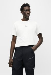 Νεαρός άνδρας που φοράει άσπρο μπλουζάκι The North Face και μαύρο παντελόνι με τσέπες με φερμουάρ, στέκεται με το ένα χέρι στην τσέπη.