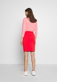 Maglietta rosa a maniche lunghe abbinata a una gonna rossa aderente, con una consistenza liscia. Indossata con sneakers bianche. Sfondo bianco uniforme.