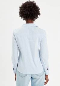 Chemise rayée bleu clair à manches longues et col pointu. Présente des poignets avec des accents de bouton et un ourlet incurvé. Fabriquée en tissu lisse.