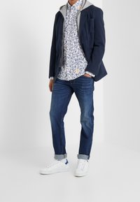 Giacca marina con cappuccio grigio, camicia bianca con stampa, jeans blu e sneaker bianche con accenti blu. Un outfit casual moderno con elementi di sovrapposizione.