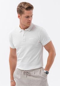 Bílá polo košile s tříknoflíkovým zapínáním, krátkými rukávy a texturovanou látkou. Spárovaná se světle šedými kalhotami na stažení a stříbrnými hodinkami.