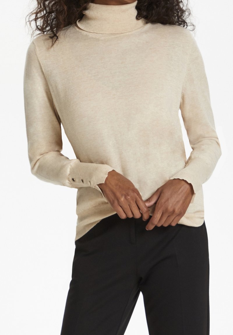 Beige coltrui met lange mouwen en knoopaccenten, geribde manchetten en een gladde, lichte textuur. Zwarte broek eronder.