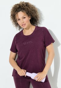 Kurzärmliges, rundhalsiges burgunderrotes T-Shirt mit "Sport"-Druck, kombiniert mit passenden Unterteilen und einer helllila Wasserflasche in der Hand.