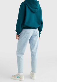 Felpa con cappuccio di colore teal, con polsini a coste, abbinata a jeans azzurri chiari con toppe ricamate e sneakers tie-dye multicolori.