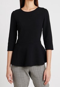 Donna che indossa un top peplum nero a trama con maniche a tre quarti e pantaloni a quadri bianco e nero, in piedi contro uno sfondo neutro.
