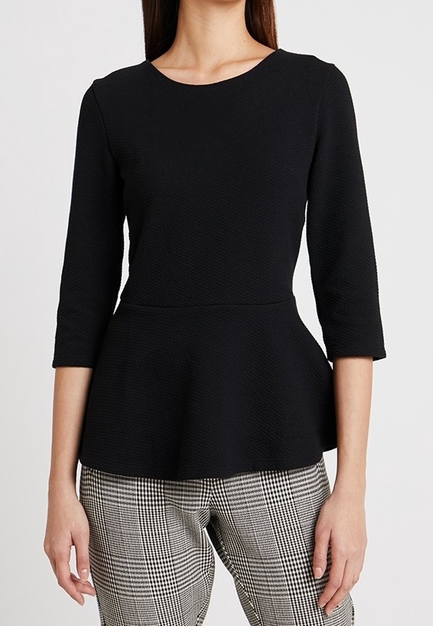 Donna che indossa un top peplum nero a trama con maniche a tre quarti e pantaloni a quadri bianco e nero, in piedi contro uno sfondo neutro.