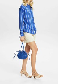 Camisa de botones a rayas azules, pantalones cortos beige, llevando un pequeño bolso azul de mimbre y sandalias de tacón nude.