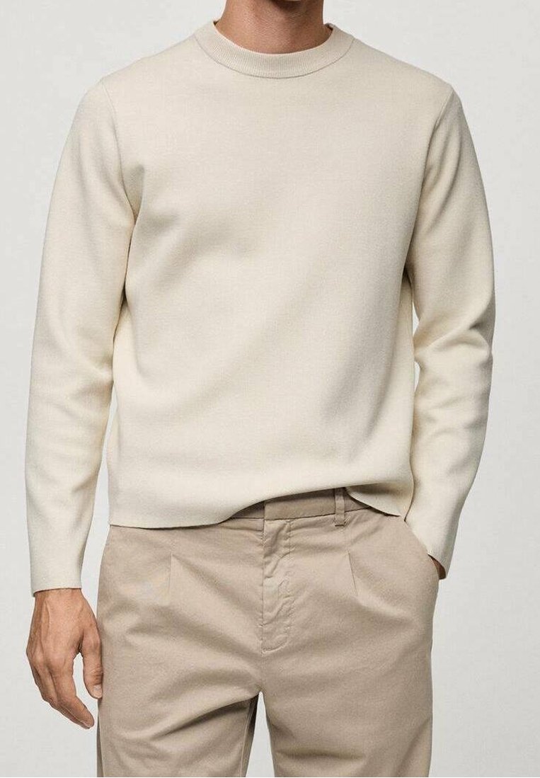 Pull beige à col rond porté avec un pantalon plissé beige clair, une main dans la poche, sur un fond neutre.