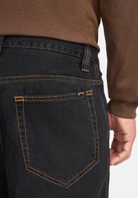 Dettaglio della tasca posteriore di jeans neri di denim con cuciture arancioni, indossati con una camicia a maniche lunghe marrone, che mostra parte di una mano sul lato.