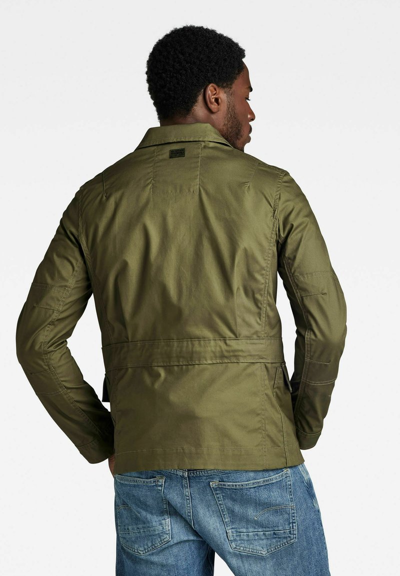 Giacca leggera di colore verde oliva con colletto strutturato, cintura in vita e cuciture visibili. Abbinata a jeans blu, che mostrano un look casual.
