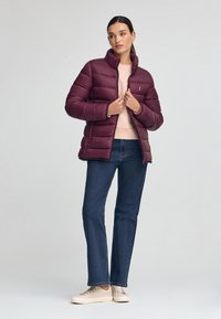 Bordeaux puffjacka med quiltat mönster, hög krage, dragkedjsstängning, bärs över en ljus tröja och blå jeans, matchad med beige sneakers.