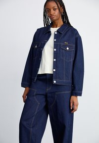 Casaco de ganga azul escuro com costuras claras, bolsos frontais e botões de pressão. Combinado com calças de ganga largas e à medida.