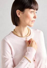 Gullkjede og armbånd, båret med en lys rosa cardigan med rund hals og knappedetaljer. Glatt tekstur, diskré design.