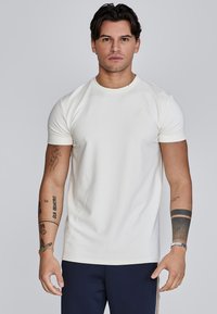 Camiseta corta de manga en color crema, confeccionada con tela suave, con cuello redondo y un discreto detalle de logo en el pecho. Llevada con pantalones navy.