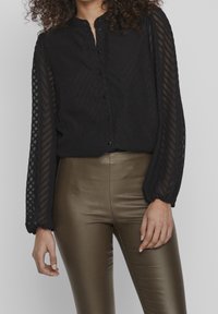 Blouse noire boutonnée avec tissu texturé, manches longues transparentes et col mandarin, assortie à un pantalon bronze ajusté et brillant.