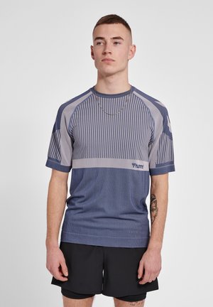 Ung mand iført en blå og grå kortærmet sportstrøje og sorte shorts, stående mod en ensfarvet lys baggrund.