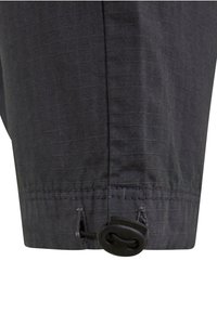 Primer plano de una tela oscura con un dobladillo cosido y un seguro para cordón de plástico negro ajustable unido a un cordón elástico fino.