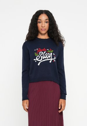 Femeie cu păr lung și creț, purtând un pulover bleumarin cu inscripția "Santa Slay" decorat cu crenguțe de ilex și o fustă plisată burgundă.