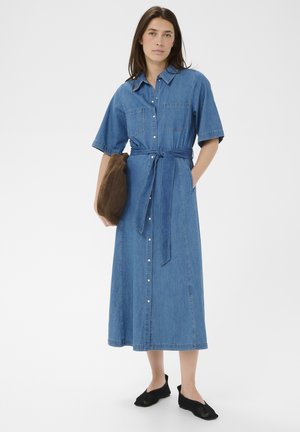 Nő kék farmer midi ruhát visel rövid ujjakkal és övvel, egy barna borítéktáskát tart, fekete hálós cipőben áll fehér háttér előtt.