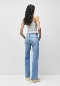 Mujer con cabello rizado y largo, vestida con una camiseta sin mangas blanca, jeans rectos azules claros y sandalias de plataforma beige, de pie contra un fondo liso.