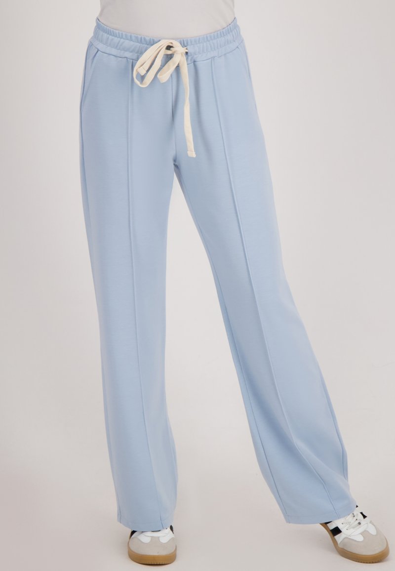 Lichtblauwe, ontspannen fit broek met voornaad, een elastische tailleband, crèmekleurige trekkoord, gedragen met witte sneakers op een neutrale achtergrond.