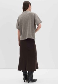 Frau steht mit dem Rücken zur Kamera, trägt ein kurzärmliges Tweedoberteil, einen langen dunklen Rock und schwarze High Heels vor einem schlichten Hintergrund.