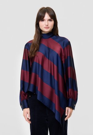 Une femme porte un chemisier à manches longues à rayures bordeaux et bleu marine, avec un ourlet asymétrique, ainsi qu’un pantalon foncé sur un fond uni.