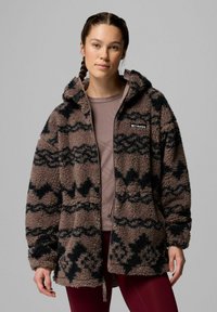 Giacca in pile marrone con un motivo a zigzag nero, chiusura con zip frontale e cappuccio. Indossata sopra una maglietta marrone chiaro, caratterizzata da una vestibilità aderente e una texture morbida.