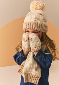 Strikket hat med pom-pom og blomsterdekorationer, parret med matchende vanter og tørklæde. Farverne inkluderer creme og beige. Tekstureret stof.