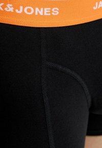 Melna sporta bikses ar oranžu elastīgu jostu un baltu logo. Gluda auduma struktūra ar plakanām šuvēm un pieguļošu siluetu.