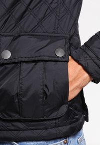 Sort kviltet jakke med en glat, skinnende overflade, metal trykknapper og en forlomma med en klapdesign.