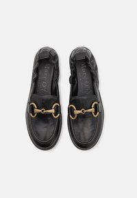 Chaussures en cuir noir sans lacets, présentant un bout rond, un bord froncé et un accent chaîne dorée proéminent sur le devant.