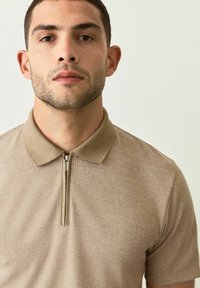 Polo de color beige con cremallera, con un patrón de cuadrícula texturizado, mangas cortas y un cuello suave. El material parece liso y ligeramente estructurado.