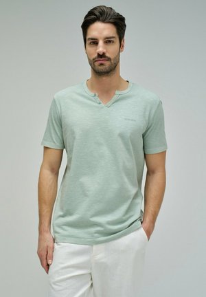 Hombre con cabello oscuro y barba, vistiendo una camiseta henley de manga corta verde menta y pantalones blancos, de pie con una mano en el bolsillo.