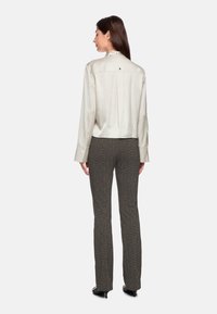 Frau, die wegschaut und eine cremefarbene Satin-Bluse mit langen Ärmeln sowie braune, strukturierte Slim-Fit-Hosen und schwarze High Heels trägt, vor weißem Hintergrund.