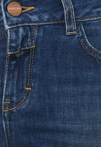 Jean en denim bleu foncé avec des coutures dorées, présentant un bouton en bronze avec "CLOSED" embossé et une petite étiquette de logo noire.