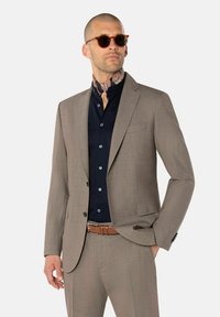 Hellbraunes, tailliertes Anzug mit passgenauer Schnittführung, kombiniert mit einem dunkelblauen Hemd mit Knöpfen. Das Modell trägt braune Sonnenbrillen und einen gemusterten Schal. Brauner Gürtel.