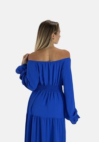 Kvinde med langt hår iført en blå off-shoulder kjole med lange ærmer, set bagfra mod en ensartet hvid baggrund.