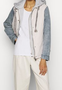 Letvægtsjakke med grå hætte, beige front og blå denimærmer, med dekorative frynser og en sølv lynlås.